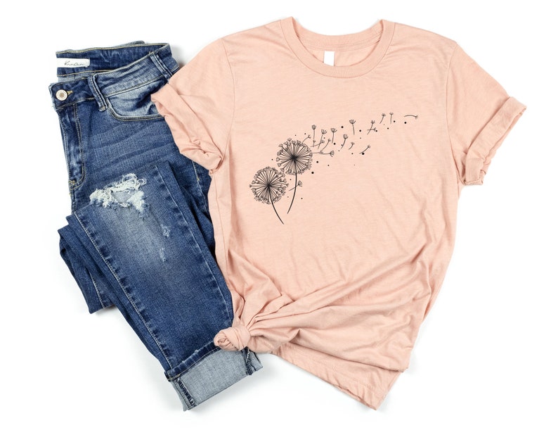 May include: A peach-colored t-shirt with a black dandelion graphic. The dandelion is blowing in the wind. The t-shirt is paired with blue jeans.