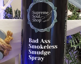 May include: A black glass bottle with a spray nozzle, labelled "Bad Ass Smokeless Smudge Spray". The bottle contains organic essential oils, including sage, rue, palo santo, hyssop, basil, and dead sea salt. The label also states that the spray is charged by full moon and Reiki master energy for clearing negative energy.
