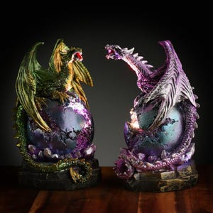 May include: Two dragon figurines, one green and one purple, are perched atop iridescent purple and green orbs. The dragons are in a fighting pose, with their wings spread and their tails curled. The orbs are surrounded by crystals and rocks, giving the figurines a magical and mystical feel.