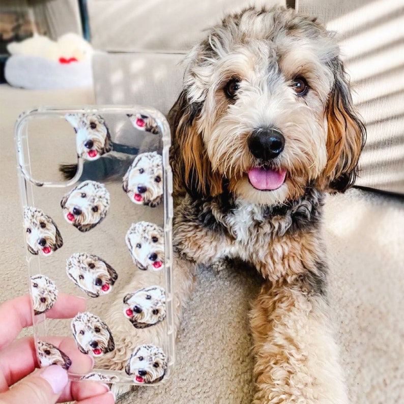 May include: A clear phone case with a repeating pattern of small dog faces. The case is being held by a hand. A brown and white dog is sitting in the background.