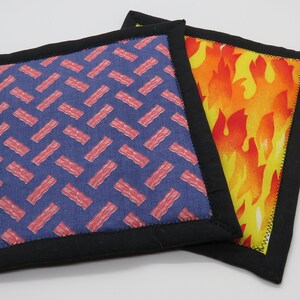 May include: Two pot holders with black trim. One pot holder has a blue background with a pink bacon pattern. The other pot holder has a yellow background with an orange flame pattern.
