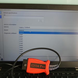 Może przedstawiać: Czarny laptop z oprogramowaniem diagnostycznym do pojazdów, wyświetlający menu wyboru modeli Legacy i Outback, oraz jaskrawopomarańczowe urządzenie diagnostyczne J2534 Pro OBDII z czarnym kablem spoczywającym na klawiaturze.