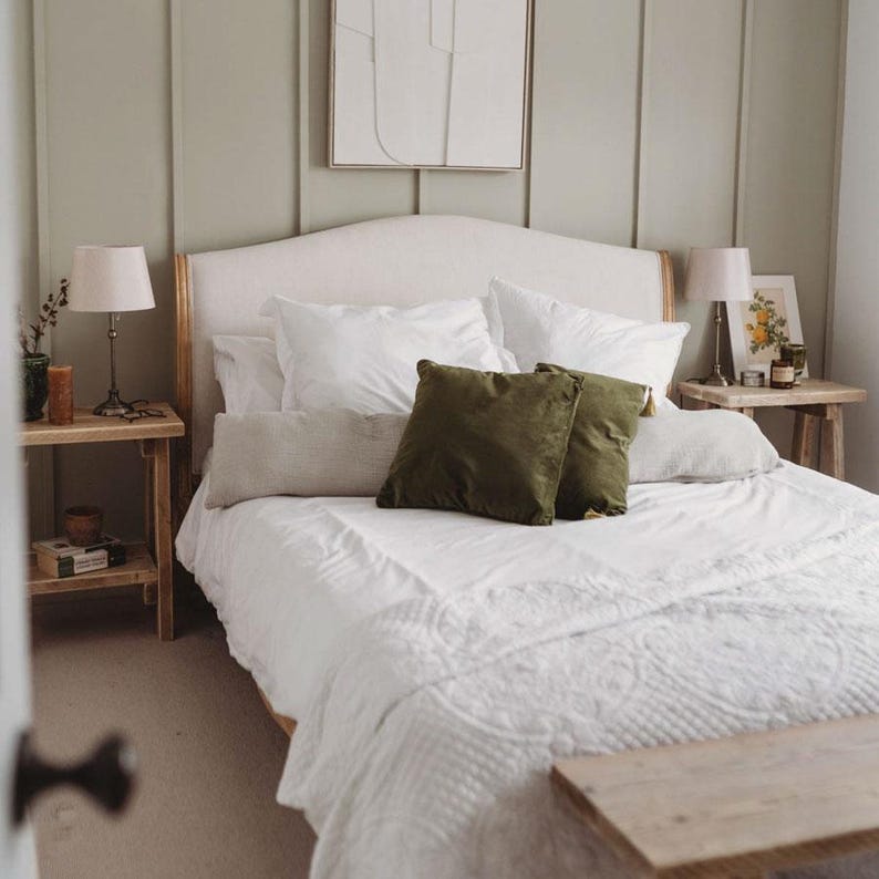 K&ouml;nnte beinhalten: Ein Bett mit einem hellen, gepolsterten Kopfteil, wei&szlig;er Bettw&auml;sche und zwei quadratischen olivgr&uuml;nen Zierkissen. Das Bett wird von zwei Holznachttischen flankiert, die jeweils eine Lampe mit wei&szlig;em Schirm tragen, vor einer salbeigr&uuml;nen Wand.