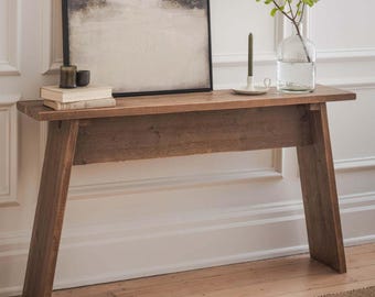 May include: A rustic, wooden console table with a natural finish. The table features a long, rectangular top and angled legs. A framed landscape painting, books, candles, and a glass vase with greenery are displayed on the table.