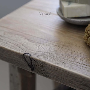 K&ouml;nnte beinhalten: Eine helle Holzoberfl&auml;che mit sichtbarer Maserung und einem dunklen Astloch an der Kante. Das Wort Natural ist in schwarzer Schrift auf das Holz gedruckt. Ein kleiner Teil einer grauen Keramikschale und ein strukturiertes beiges Objekt sind sichtbar.
