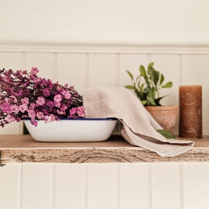 May include: A wooden shelf holds a white enamel basin with a blue rim, filled with pink flowers and a beige linen cloth. Two brown pillar candles and a potted plant are also present. The shelf is made of reclaimed wood.