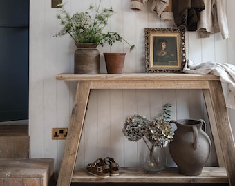 May include: A rustic wooden console table with a lower shelf. The table has a distressed finish and is decorated with a vase of white flowers, a potted plant, a framed portrait, and a vase of hydrangeas.