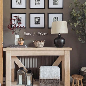 May include: A light wood console table with a shelf, featuring a white throw blanket in a basket, two black lanterns, and a lamp with a cream shade. The table is 120cm long and has a sand finish. The wall behind the table is a dark brown colour and features black framed photos.