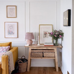 May include: A wooden console table with two woven baskets underneath. The table has a pink lamp with a fringed shade, a white candle, and a vase of pink flowers on top. The table is in a living room with a yellow sofa and a rug on the floor.