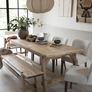 May include: A rustic wooden dining table with a bench and chairs. The table is made of natural wood and has a distressed finish. The chairs are upholstered in white fabric. The table is set with plates, bowls, and a jug.
