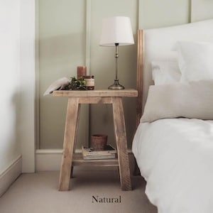 May include: A natural wood bedside table with a single shelf. The table has a lamp, a small potted plant, and a stack of books on it. The table is next to a white bed with white bedding.