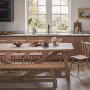 May include: A light wood farmhouse table with a bench and chairs. The table is set with a bowl of fruit, a loaf of bread, and a jar of honey. The table is in a kitchen with a window overlooking a garden.