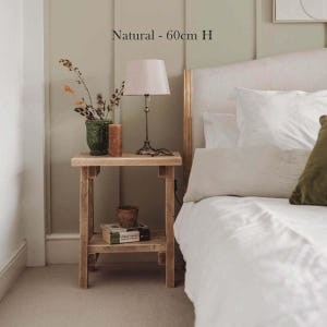 K&ouml;nnte beinhalten: Ein heller Holznachttisch mit einer unteren Ablage, der 60 cm hoch ist. Auf dem Tisch stehen eine Lampe mit wei&szlig;em Schirm, eine gr&uuml;ne, strukturierte Vase mit getrockneten Zweigen und eine braune Kerze. Ein Buch liegt auf der unteren Ablage.