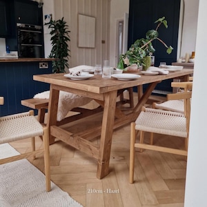 May include: A rustic wooden dining table with a natural finish and a farmhouse style. The table is 210cm long and is made of hazel wood. The table is set with white plates, glasses, and a green plant. The table is surrounded by chairs with woven seats.