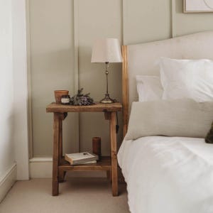 May include: A wooden bedside table with a lamp, a book, and a small potted plant. The table has two shelves and is made of reclaimed wood. The lamp has a white shade and a gold base. The book is on the lower shelf and the plant is on the upper shelf.