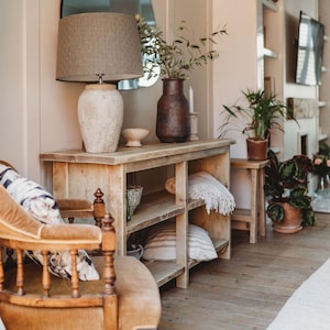 May include: A rustic wooden console table with three shelves, featuring a lamp with a beige lampshade and a brown ceramic vase with greenery. The table is decorated with throw blankets and decorative items.