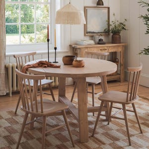 May include: A light wood round dining table with four matching chairs. The table is surrounded by a patterned rug and set near a window. A woven lampshade hangs above the table, and a side table with decor is visible in the background.