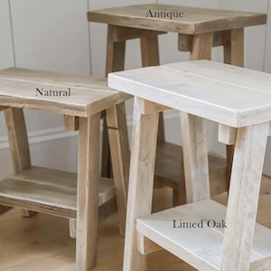 K&ouml;nnte beinhalten: Drei Holzhocker mit vier Beinen und einer unteren Ablage. Der Hocker hinten ist mit Antique beschriftet, der linke mit Natural und der vordere rechte mit Limed Oak, was drei verschiedene Holzoberfl&auml;chen und Farbt&ouml;ne zeigt.