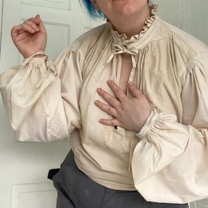 May include: A cream-coloured, long-sleeved blouse with a ruffled collar and cuffs. The blouse has a keyhole neckline with a tie closure and voluminous sleeves gathered at the wrists. The fabric appears to be lightweight and slightly textured.