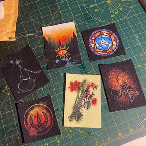 May include: Six different embroidered patches on a green cutting mat. The patches feature a variety of designs, including a sunset scene with an eye, a stained glass d20, a sword and flowers, a fire and dice, and a logo with a spear and a wreath.