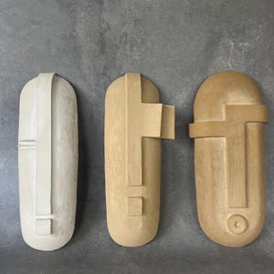 May include: Three abstract wall masks in neutral tones. The mask on the left is off-white, while the other two are shades of beige and tan. Each mask has a unique geometric design, with rectangular and cross-like features.