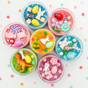 May include: A top-down view of seven small, clear jars filled with colorful, textured slime. Each jar is decorated with various miniature figurines, including Easter eggs, bunnies, a dinosaur, and a ladybug. The jars are arranged in a circular pattern.