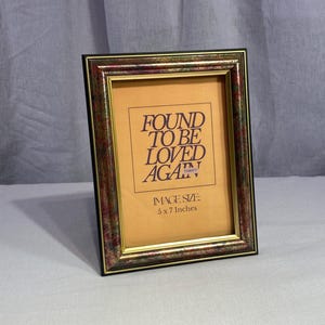 May include: A rectangular picture frame with a dark, multi-colored mottled border and a gold inner trim. The insert displays the text FOUND TO BE LOVED AGAIN and IMAGE SIZE: 5 x 7 Inches in dark lettering on a light tan background.