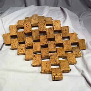 May include: A diamond-shaped trivet made of rectangular cork pieces. The cork pieces are light brown with dark brown speckles and are connected with small metal rings. The trivet is designed to protect surfaces from hot items.