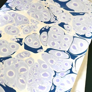 May include: A sheet of marbled paper with a cream-colored background and a pattern of blue and lavender swirls. The paper is folded, revealing a plain white sheet underneath. The overall effect is artistic and decorative.