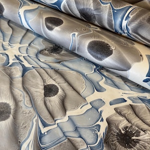 May include: Close-up of marbled paper in shades of gray, blue, and cream. The paper has a swirling pattern with dark gray circular shapes and blue veins. The paper is folded, showing multiple layers of the design.