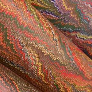 May include: A sheet of paper featuring a traditional marbled pattern with repeating wave-like designs. The surface displays a dense, intricate arrangement of vibrant orange, yellow, olive green, and white pigments, creating a fluid, multi-colored texture.
