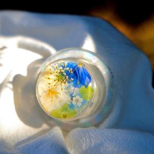 May include: A clear glass bottle stopper with a floral design. The stopper features a mix of blue, white, and yellow dried flowers encased in clear resin. The stopper is sitting on a white fabric. The text "Kristin ABLOOM Floral Design" is visible.