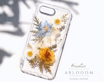 Könnte beinhalten: Eine transparente Handyhülle mit gepressten Blumen darin. Die Blumen sind weiß, blau und gelb. Die Hülle hat einen erhöhten Rand um das Kameraobjektiv. Der Text "Kristin ABLOOOOM FLORAL DESIGN" ist auf der Unterseite der Hülle aufgedruckt.