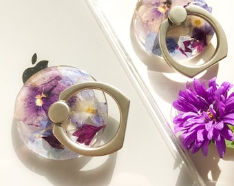 Könnte beinhalten: Zwei Handy-Ringhalter mit einem silbernen Metallring und einer durchsichtigen Harzbasis, gefüllt mit gepressten lila und blauen Blumen.