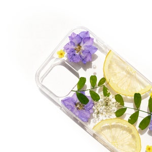 Puede incluir: Una funda transparente para tel&eacute;fono con flores prensadas y rodajas de lim&oacute;n. Las flores son moradas y amarillas, y las rodajas de lim&oacute;n son amarillas. La funda est&aacute; dise&ntilde;ada para adaptarse a un tel&eacute;fono inteligente.