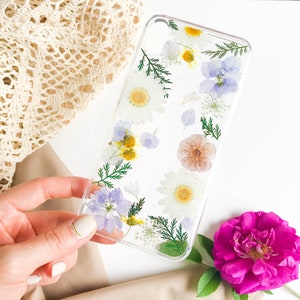 May include: A clear phone case with pressed flowers and leaves. The flowers are white, yellow, blue, and pink. The case is on a white surface.