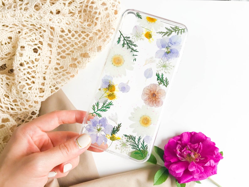 May include: A clear phone case with pressed flowers and leaves. The flowers are white, yellow, blue, and pink. The case is on a white surface.