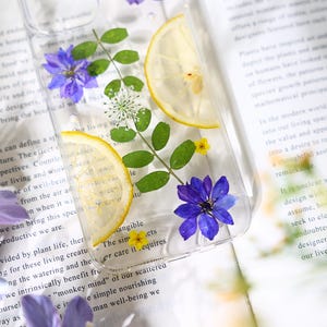 Puede incluir: Una funda transparente para tel&eacute;fono con flores prensadas y rodajas de lim&oacute;n. Las flores son moradas y blancas, y las rodajas de lim&oacute;n son amarillas. La funda est&aacute; sobre un libro con texto.