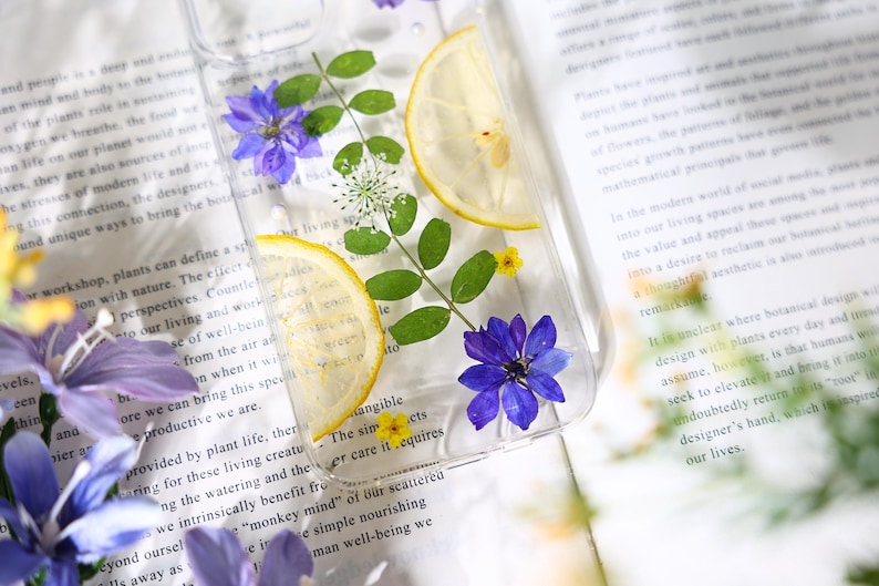 Puede incluir: Una funda transparente para tel&eacute;fono con flores prensadas y rodajas de lim&oacute;n. Las flores son moradas y blancas, y las rodajas de lim&oacute;n son amarillas. La funda est&aacute; sobre un libro con texto.