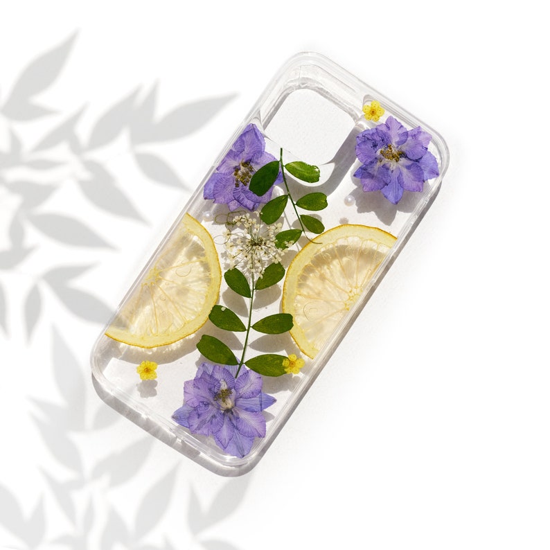 Puede incluir: Una funda transparente para tel&eacute;fono con flores prensadas y rodajas de lim&oacute;n. Las flores son moradas y amarillas, y las rodajas de lim&oacute;n son amarillas. La funda est&aacute; dise&ntilde;ada para adaptarse a un tel&eacute;fono inteligente.