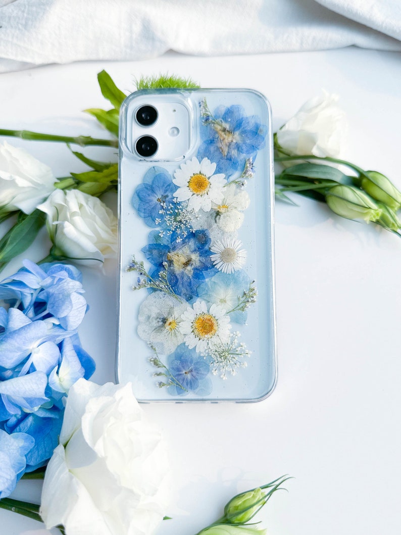 May include: A clear phone case with a design of pressed blue and white flowers. The case is on a white surface.
