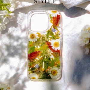 May include: A clear phone case with pressed flowers and leaves. The case features a variety of flowers, including white daisies, yellow flowers, and red leaves. The text "STYLE F" is printed on the white background behind the case.