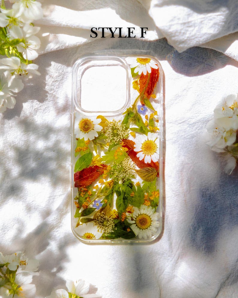 May include: A clear phone case with pressed flowers and leaves. The case features a variety of flowers, including white daisies, yellow flowers, and red leaves. The text "STYLE F" is printed on the white background behind the case.