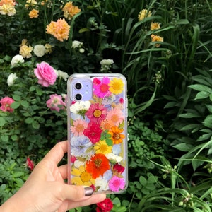 May include: A clear phone case with a design of pressed flowers in various colors, including pink, yellow, orange, and blue. The case is held in a hand.
