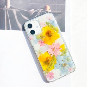 May include: A clear phone case with pressed flowers. The case features yellow, pink, and blue flowers with gold accents.