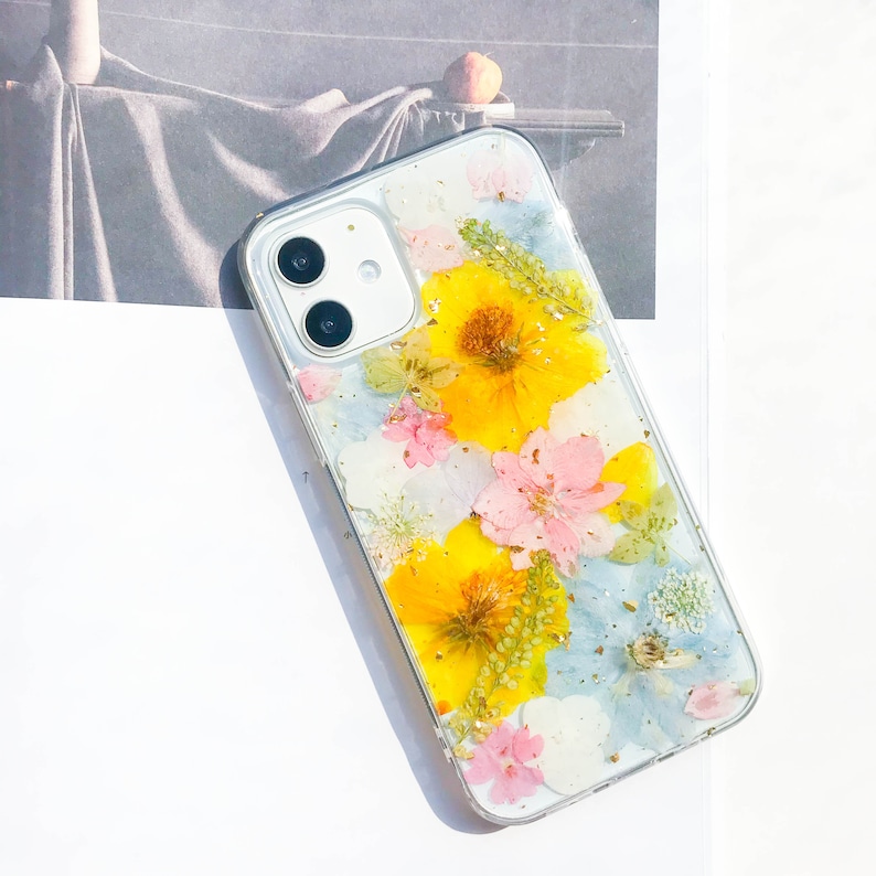 May include: A clear phone case with pressed flowers. The case features yellow, pink, and blue flowers with gold accents.