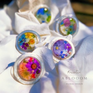 May include: Five clear resin rings with pressed flowers inside. The rings are on a white fabric background. The rings feature different flower arrangements with colours like purple, blue, yellow, and pink. The text "Kristin ABLOOOOM FLORAL DESIGN" is visible in the bottom right corner of the image.