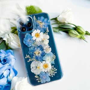 May include: A clear phone case with a teal blue background and pressed flowers. The flowers are white and blue, and they are arranged in a vertical line down the center of the case.