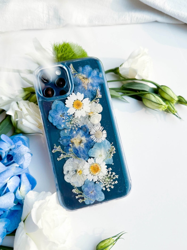 May include: A clear phone case with a teal blue background and pressed flowers. The flowers are white and blue, and they are arranged in a vertical line down the center of the case.