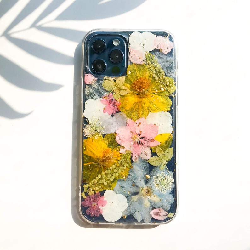 May include: A clear phone case with a pressed flower design. The flowers are yellow, pink, white, and blue. The case is made of a clear material and has a black background.